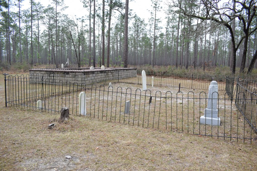 Historic gravestones and family burial plots enclosed by black iron fencing and low stone walls at Columbus Cemetery, surrounded by tall pine trees in Suwannee River State Park, Florida.