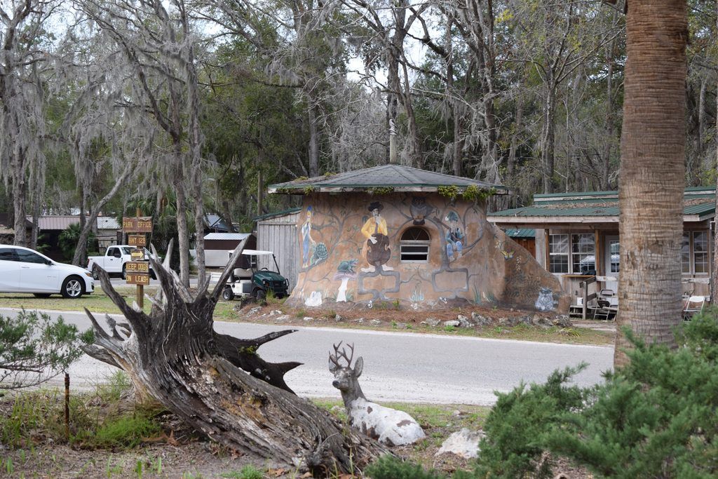 Rustic campground buildings and a round, mural-covered structure at Spirit of the Suwannee Music Park in Live Oak, Florida, surrounded by moss-draped trees and a decorative deer sculpture.