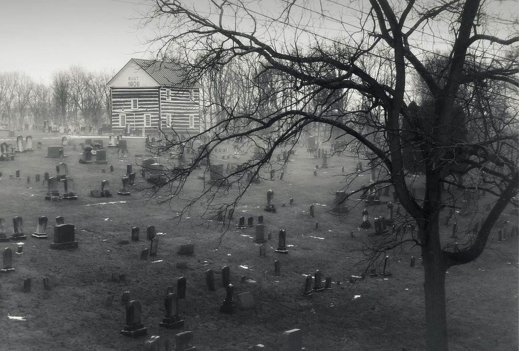 Black-and-white photograph of the 1806 Old Log Church overlooking rows of historic gravestones in Schellsburg, Pennsylvania, with a large leafless tree in the foreground.