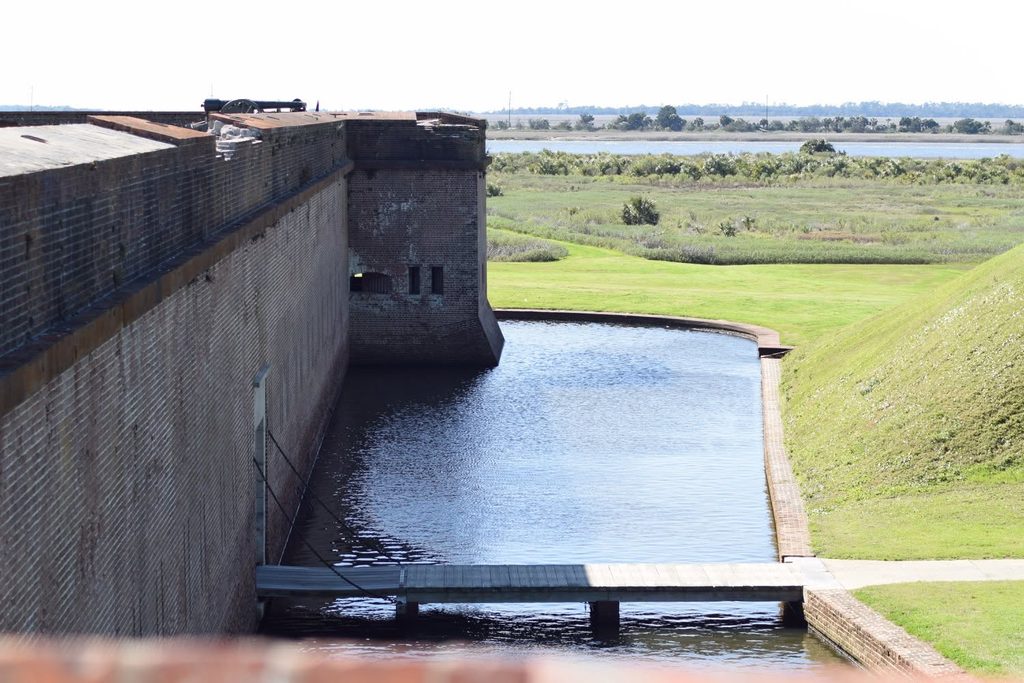 Historic brick walls, defensive moat, and wooden footbridge at Fort Pulaski National Monument in Georgia, with coastal marshlands and the Savannah River area visible beyond the fort.