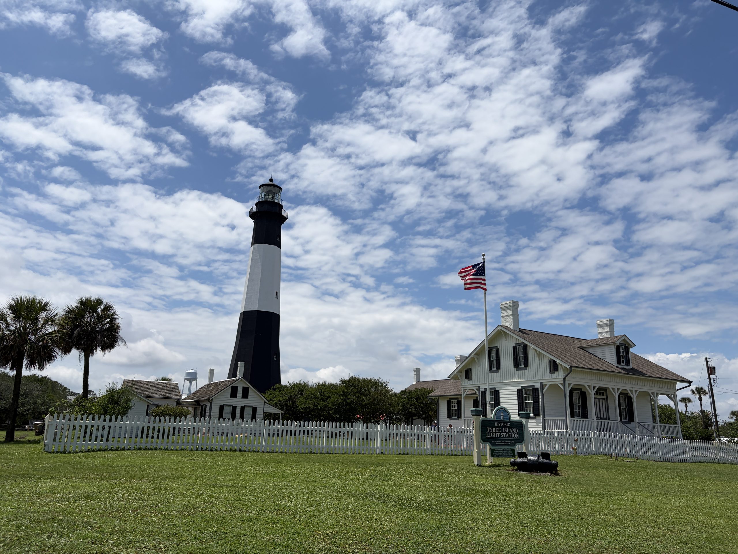 Historic black-and-white Tybee Island Lighthouse standing beside the white keeper's houses, American flag, and picket fence beneath a partly cloudy blue sky on Tybee Island, Georgia.