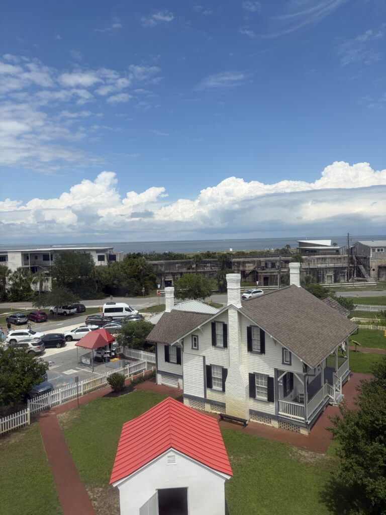 Elevated view from Tybee Island Lighthouse overlooking a historic white keeper's house, red-roofed outbuilding, Fort Screven's concrete fortifications, and the Atlantic Ocean beneath a blue sky filled with clouds.