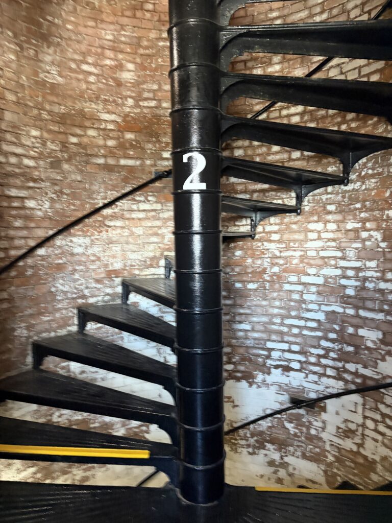Black metal spiral staircase winding around a central column marked with the number two inside Tybee Island Lighthouse, surrounded by historic exposed brick walls.
