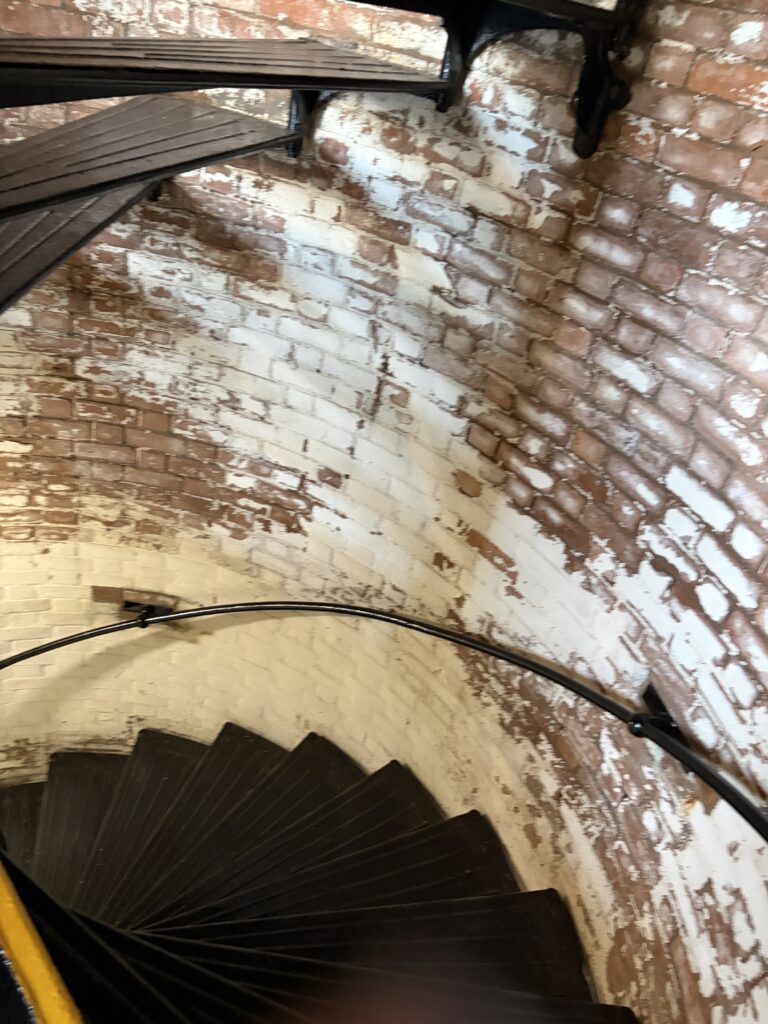 View looking down the narrow black spiral staircase inside Tybee Island Lighthouse, with curved brick walls, weathered white paint, and a metal handrail.