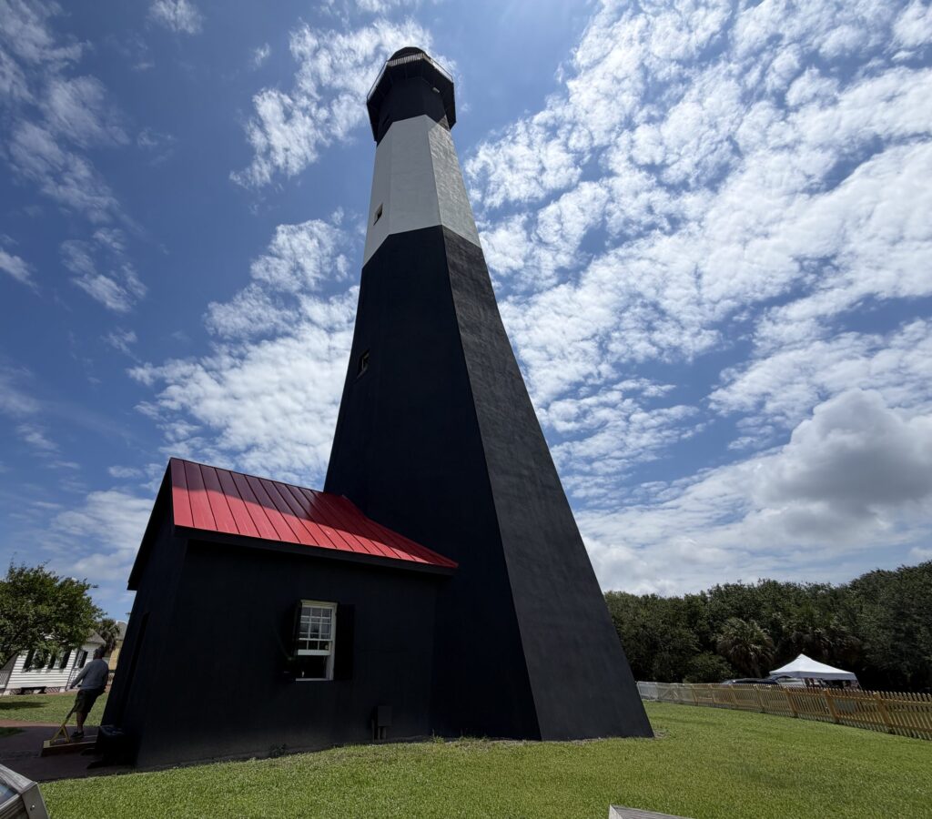 Dramatic upward view of the black-and-white Tybee Island Lighthouse in Georgia, with its red-roofed entrance building beneath a blue sky filled with scattered clouds.