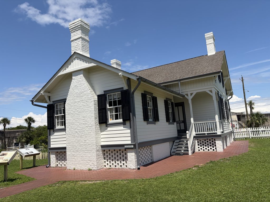 Restored white wooden lighthouse keeper's house at Tybee Island Light Station, featuring black shutters, brick chimneys, a covered porch, and entrance steps beneath a clear blue Georgia sky.