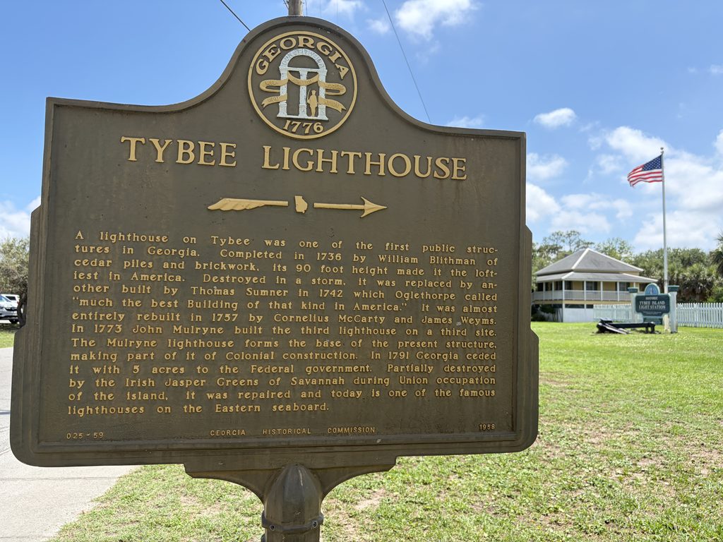 Georgia historical marker titled โTybee Lighthouse,โ detailing the lighthouse's construction in 1736, subsequent rebuilding, and Civil War damage, with the historic light station grounds visible in the background.