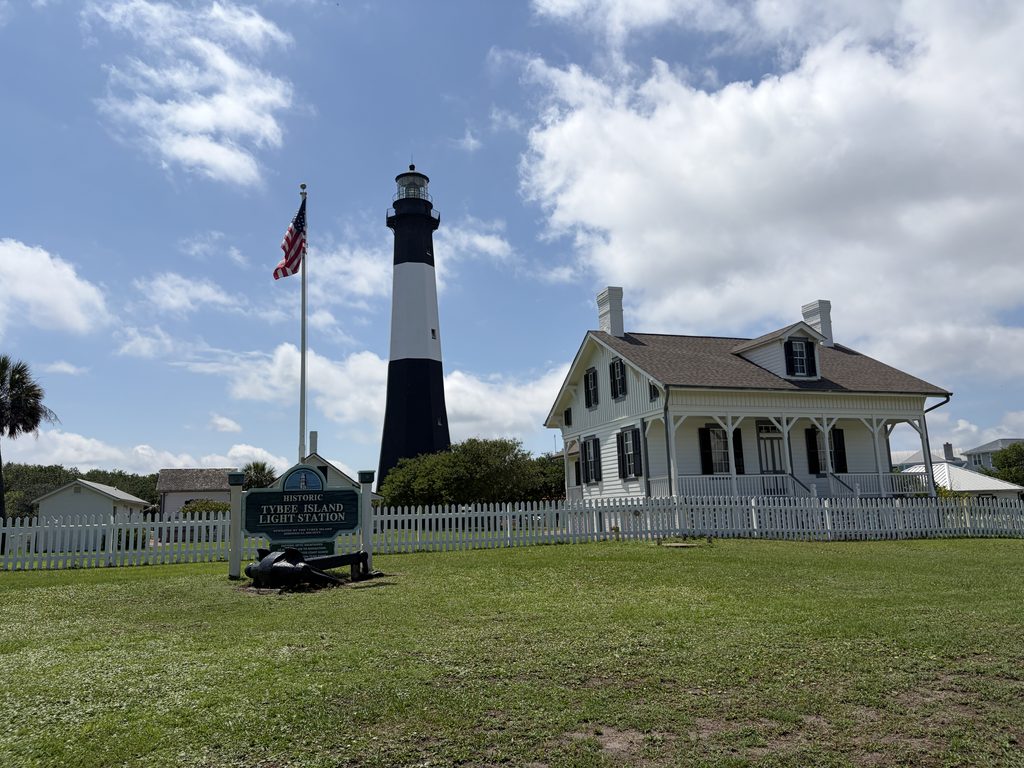 Historic Tybee Island Lighthouse and white keeper's house behind a picket fence, with the light station entrance sign, American flag, and green lawn beneath a partly cloudy sky.