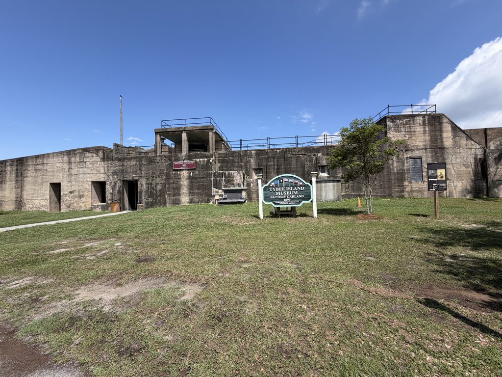 Historic concrete coastal artillery fortification at Battery Garland, part of Fort Screven on Tybee Island, Georgia, featuring the Tybee Island Museum entrance sign, weathered defensive walls, and elevated gun positions beneath a blue sky.