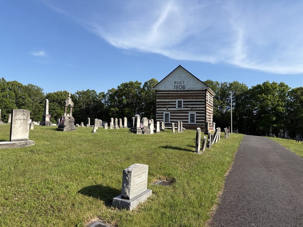 Historic 1806 Old Log Church surrounded by weathered gravestones at Chestnut Ridge and Schellsburg Union Cemetery in Bedford County, Pennsylvania, beneath a clear blue sky.