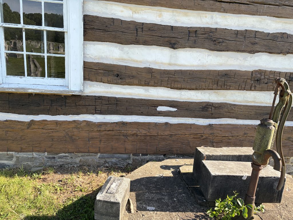 Rusted antique hand water pump beside the log wall and white-framed window of the 1806 Old Log Church in Schellsburg, Pennsylvania, with historic cemetery gravestones reflected in the glass.