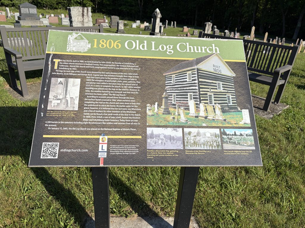 Interpretive historical marker for the 1806 Old Log Church in Schellsburg, Pennsylvania, featuring photographs, church history, and preservation information, with cemetery gravestones and benches in the background.