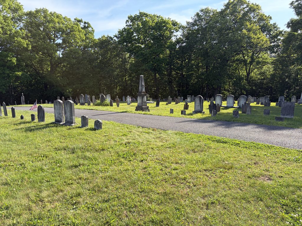 Peaceful view of historic gravestones and a tall stone monument beside a winding cemetery road at Chestnut Ridge and Schellsburg Union Cemetery in Pennsylvania, surrounded by sunlit grass and mature trees.
