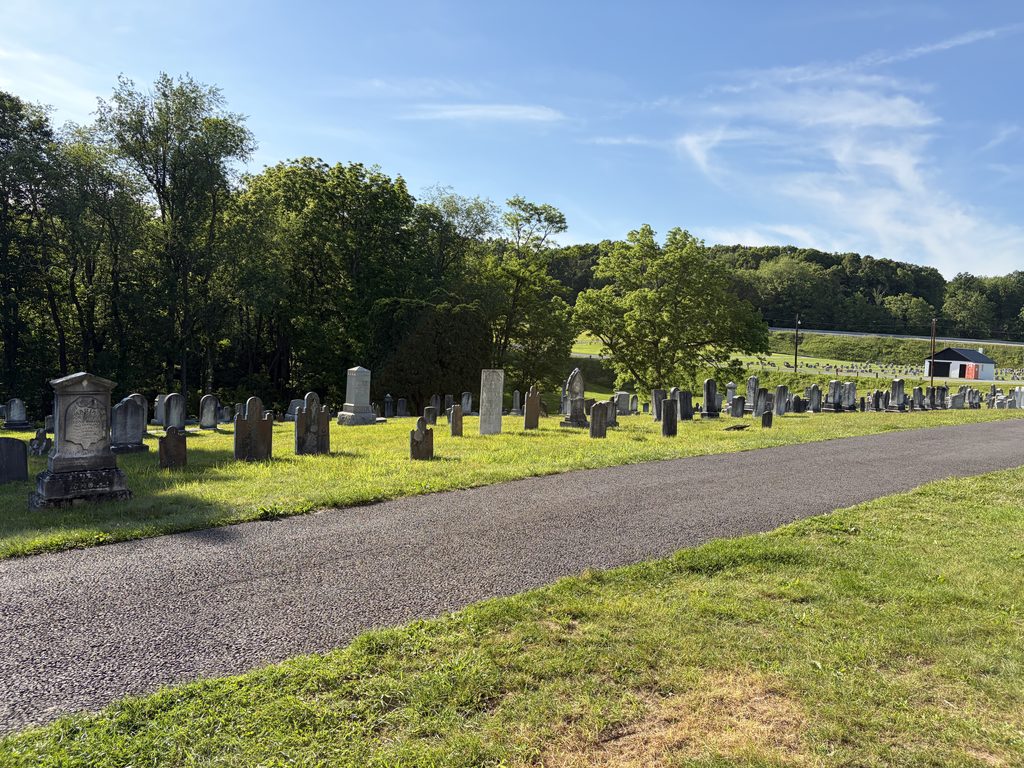 Paved cemetery road winding past historic gravestones at Chestnut Ridge and Schellsburg Union Cemetery in Schellsburg, Pennsylvania, with green trees, rolling hills, and a blue sky.