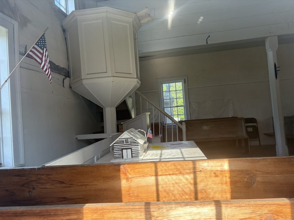 Interior of the 1806 Old Log Church in Schellsburg, Pennsylvania, showing its elevated white wineglass-shaped pulpit, wooden pews, staircase, upper gallery, and a miniature model of the church.