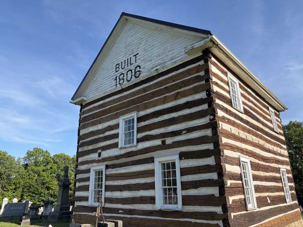 Close-up of the 1806 Old Log Church in Schellsburg, Pennsylvania, showing its exposed hand-hewn logs, white chinking, wooden windows, and “Built 1806” inscription beneath the roof.