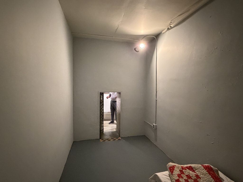 Interior of a historic jail cell at the Old Franklin County Jail in Chambersburg, Pennsylvania, showing a four-foot-tall doorway, gray walls, and a narrow bed with a quilt.