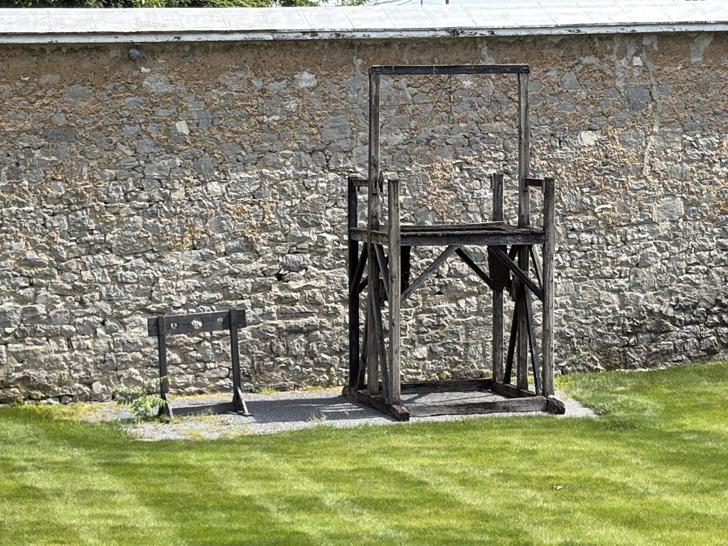 Wooden gallows with a hanging noose standing beside a smaller restraint structure in the grassy courtyard of the Old Franklin County Jail, against a tall limestone prison wall.