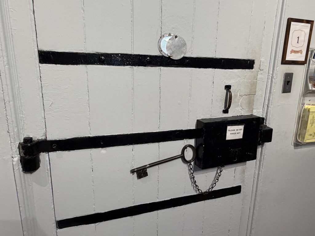 Massive white wooden jail door reinforced with black iron bars, featuring a large skeleton key chained to its lock at the Old Franklin County Jail in Chambersburg, Pennsylvania.