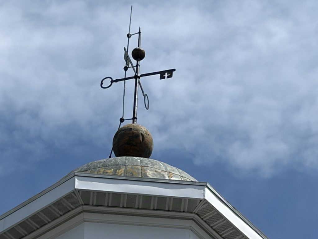 Historic weather vane shaped like two crossed skeleton keys rising above the cupola of the Old Franklin County Jail in Chambersburg, Pennsylvania, against a cloudy blue sky.