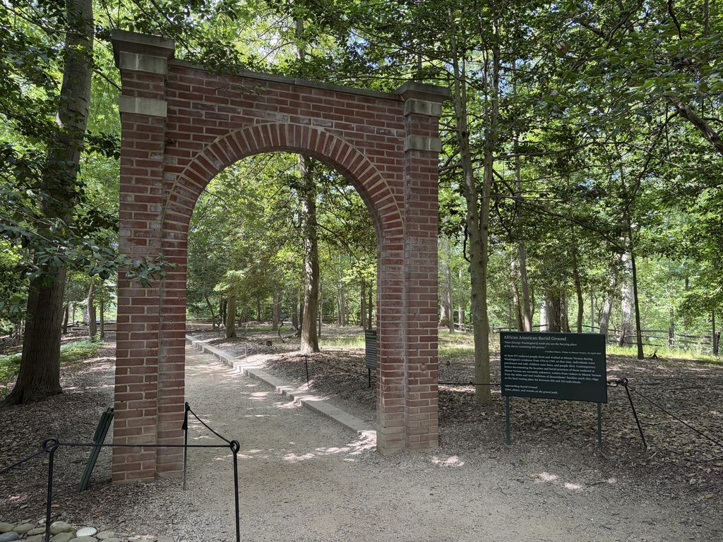 Red brick archway marking the entrance to the African American Burial Ground at Mount Vernon, Virginia, with an informational sign and wooded walking path beyond.