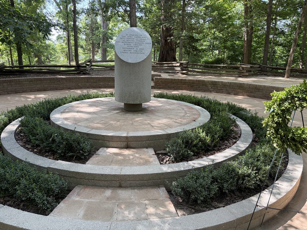Stone memorial honoring enslaved people at George Washington’s Mount Vernon Estate in Virginia, surrounded by circular brick pathways, landscaped greenery, and a wooded burial ground.
