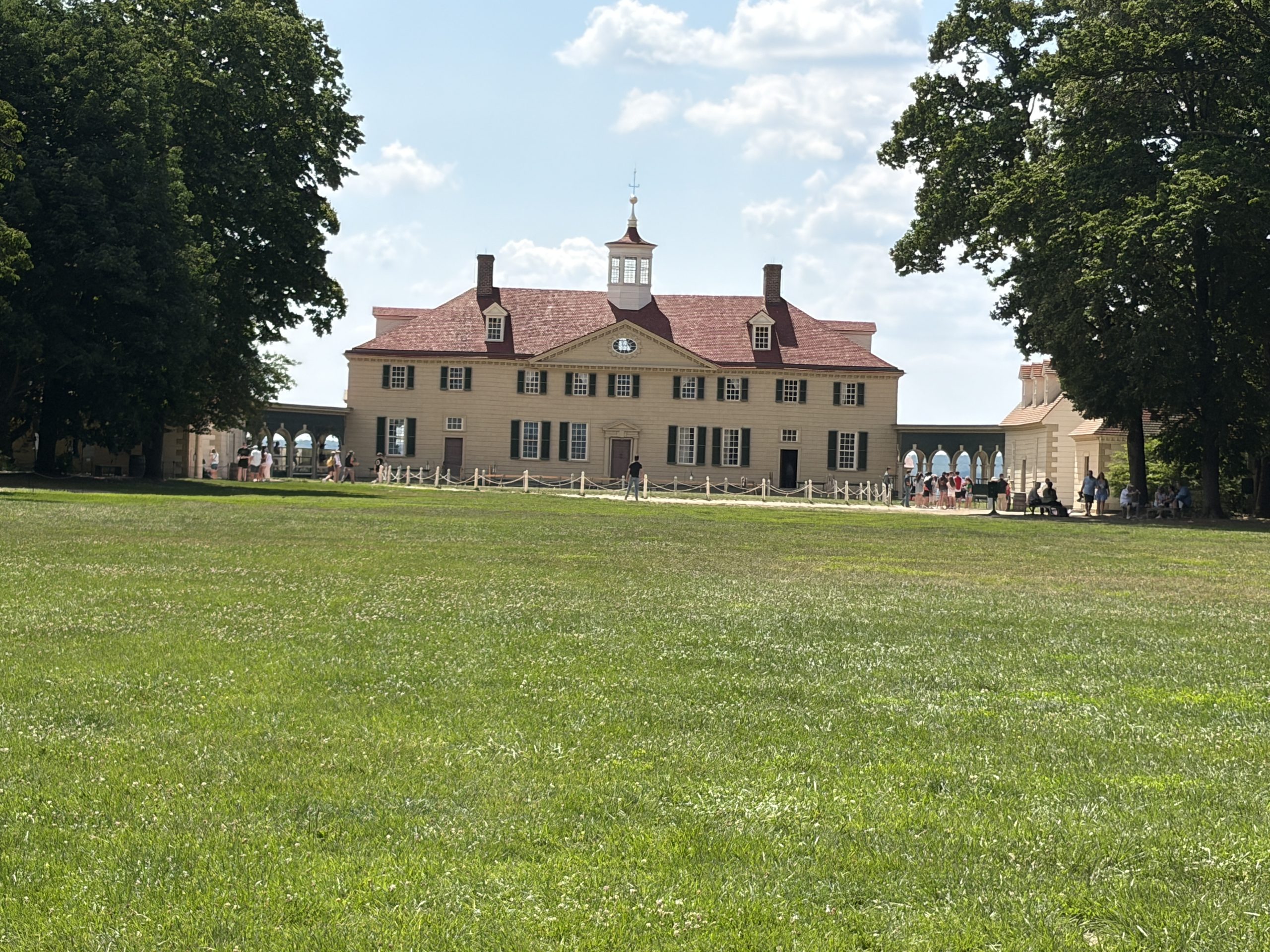 Front view of George Washington’s historic Mount Vernon mansion in Virginia, framed by large trees with visitors exploring the grounds beneath a blue sky.
