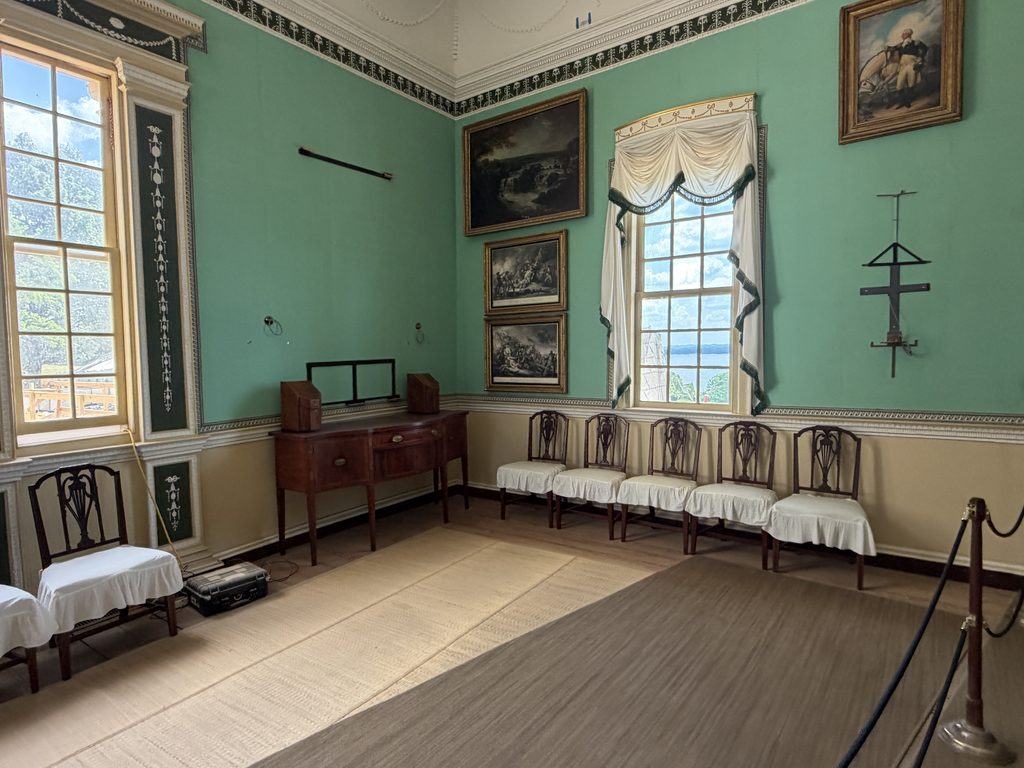 Historic room inside George Washington’s Mount Vernon mansion featuring mint-green walls, ornate trim, framed artwork, antique wooden furniture, and a row of chairs beneath a window.