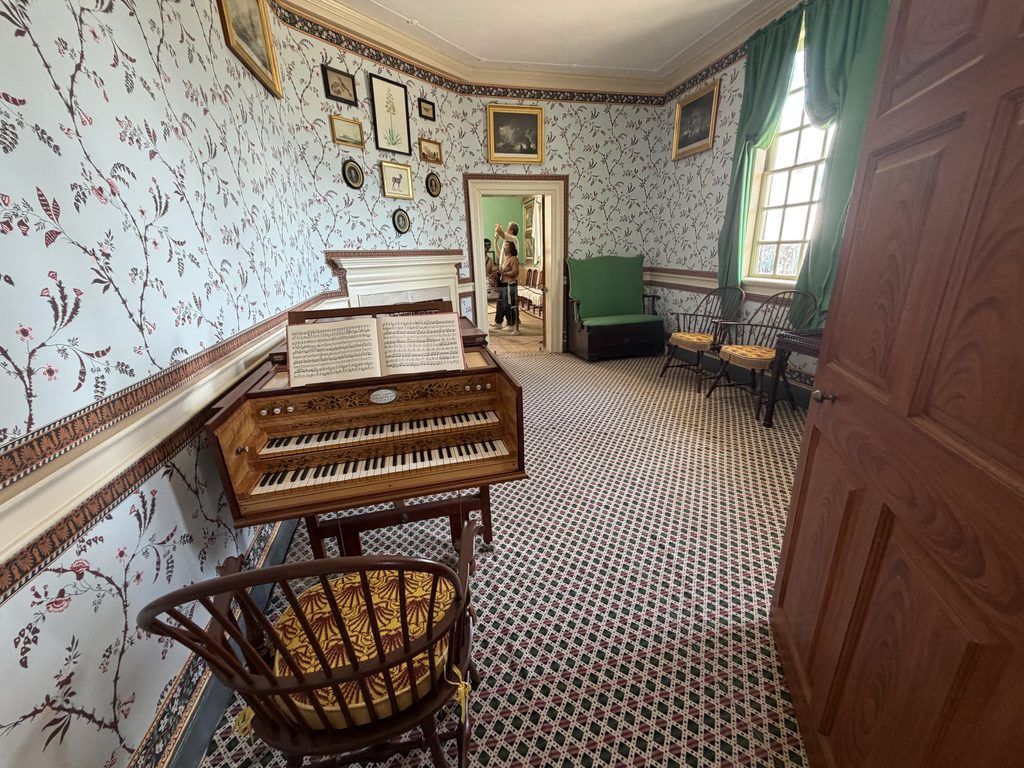 Historic Little Parlor inside George Washington’s Mount Vernon mansion, featuring patterned wallpaper, an antique keyboard instrument, framed artwork, wooden chairs, and green window curtains.