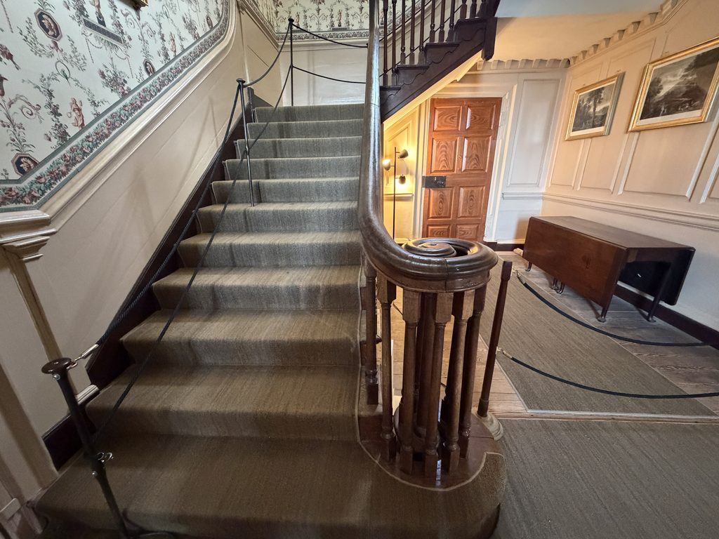 Carpeted wooden staircase inside George Washington’s Mount Vernon mansion, featuring a curved banister, decorative wallpaper, and a paneled wooden door beneath the stairs.