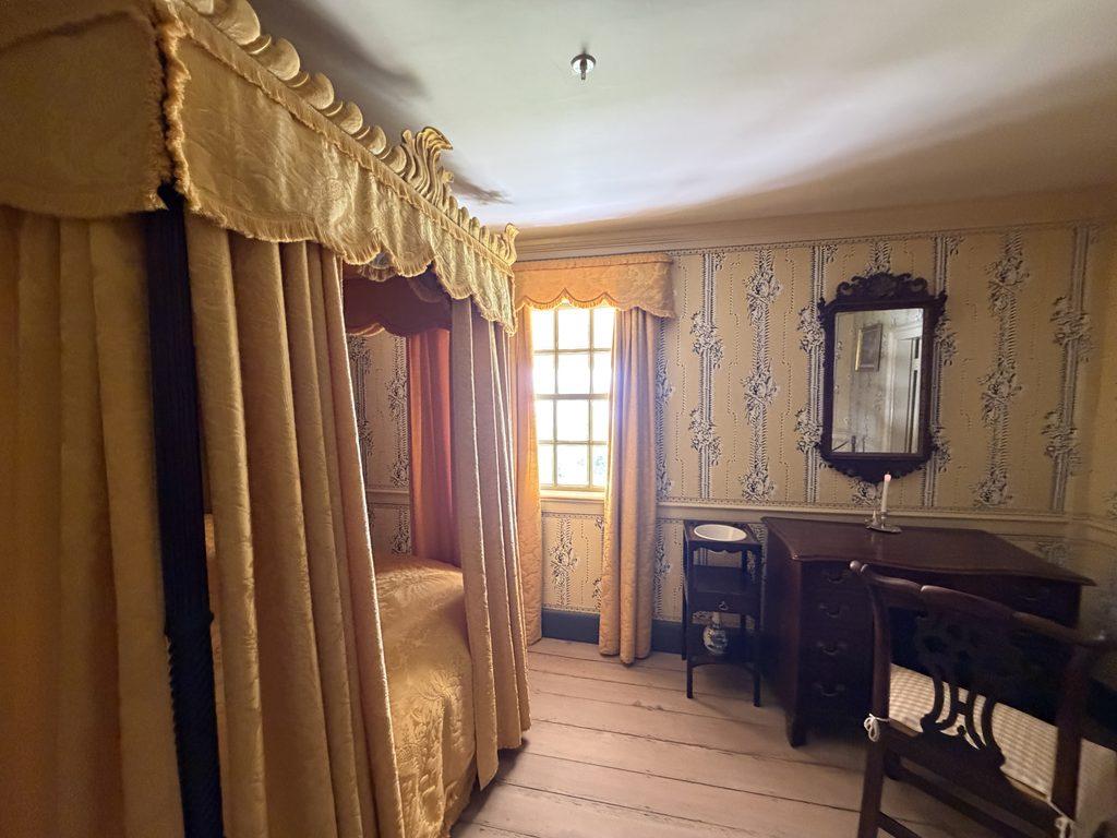 Historic Yellow Room inside George Washington’s Mount Vernon mansion, featuring a gold-curtained canopy bed, patterned wallpaper, wooden floors, an antique writing desk, and a wall mirror.