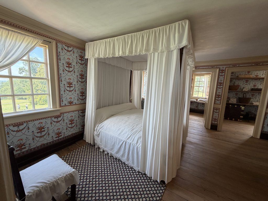 George Washington’s historic bedchamber at Mount Vernon, featuring a white-curtained canopy bed, patterned wallpaper, wooden floors, and windows overlooking the estate grounds.