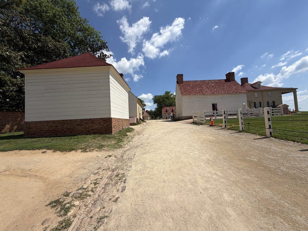 Gravel pathway winding between white wooden outbuildings with red roofs at George Washington’s Mount Vernon Estate in Virginia, with the historic mansion visible on the right beneath a bright blue sky.