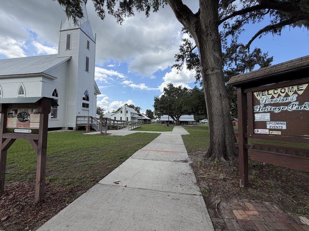 Walking path through Homeland Heritage Park in Polk County, Florida, with the historic white Methodist church, welcome sign, large oak tree, and preserved pioneer buildings in the background.