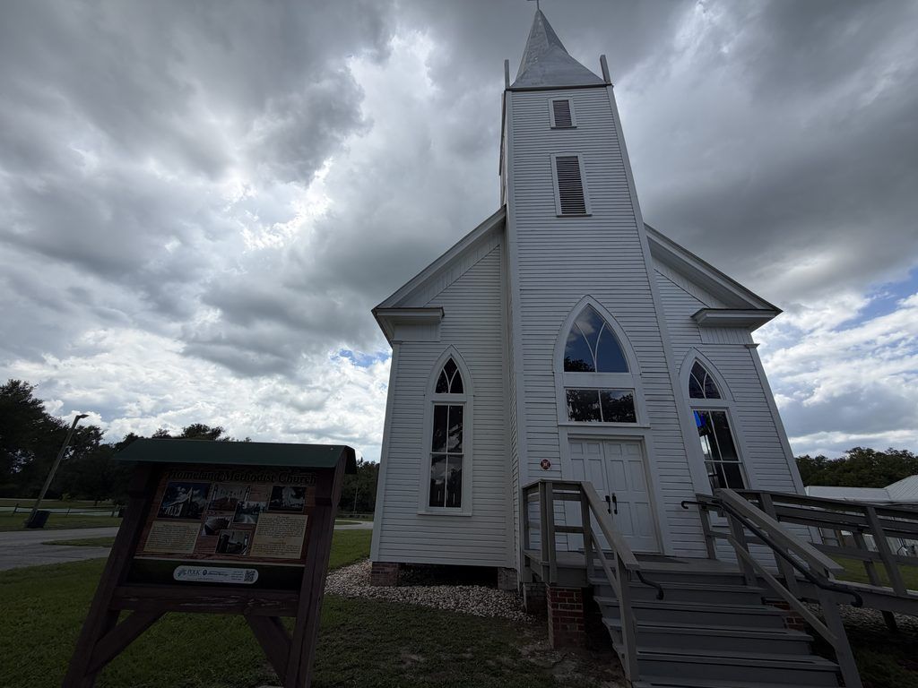 Historic white wooden Homeland Methodist Church with a tall steeple and Gothic-style windows beneath dramatic cloudy skies at Homeland Heritage Park in Polk County, Florida.