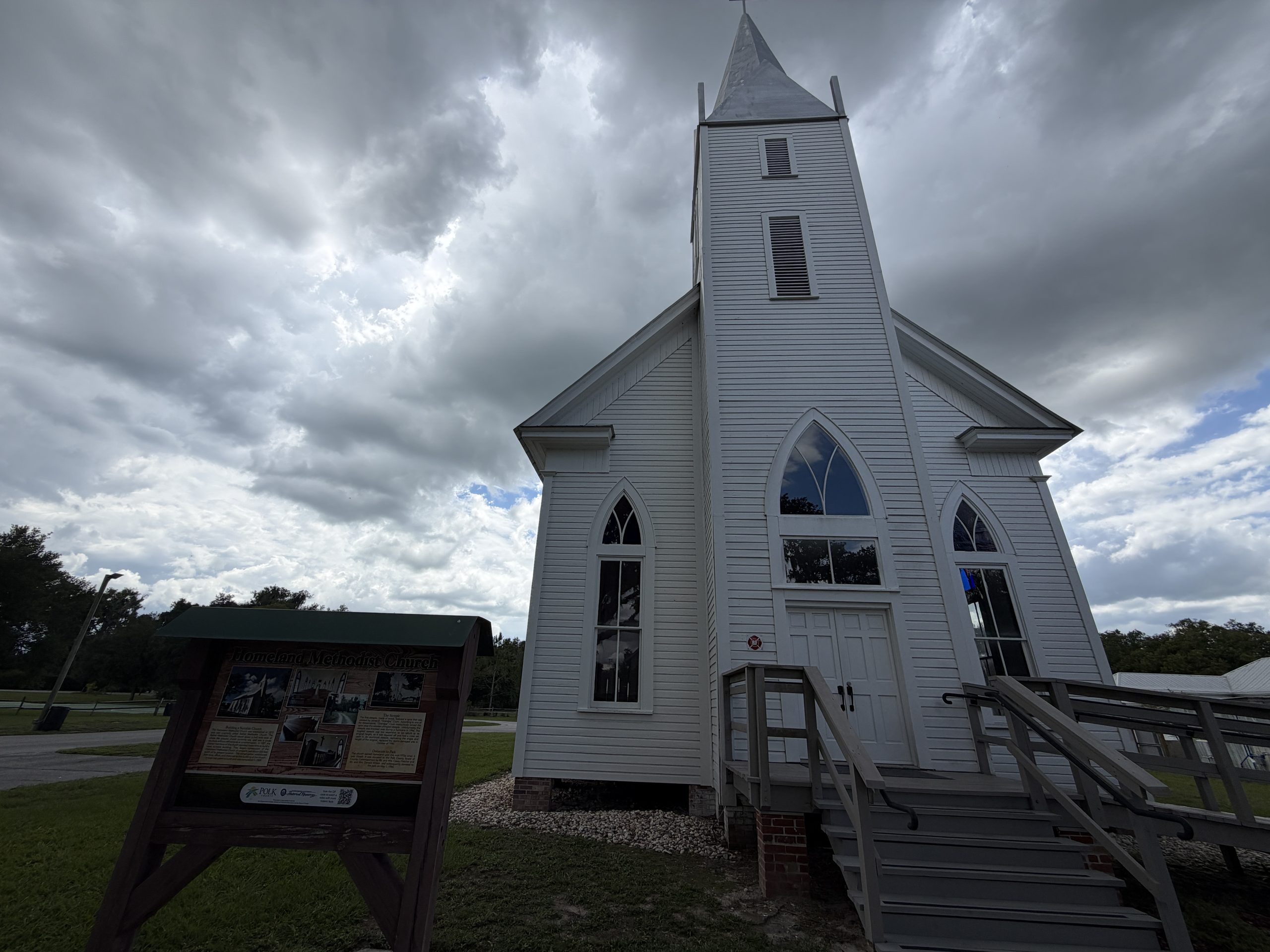 Historic white wooden Homeland Methodist Church with a tall steeple and Gothic-style windows beneath dramatic cloudy skies at Homeland Heritage Park in Polk County, Florida.