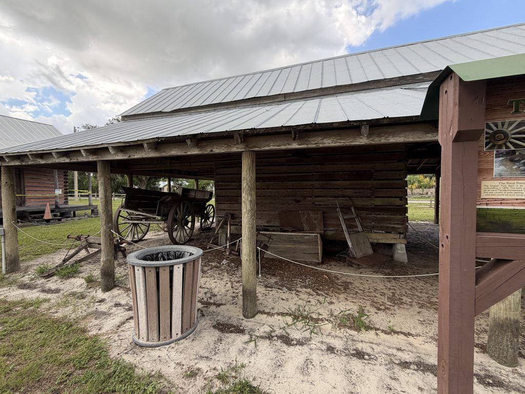 Historic wooden pole barn at Homeland Heritage Park in Polk County, Florida, sheltering an antique wooden farm wagon and agricultural equipment beneath a metal roof.