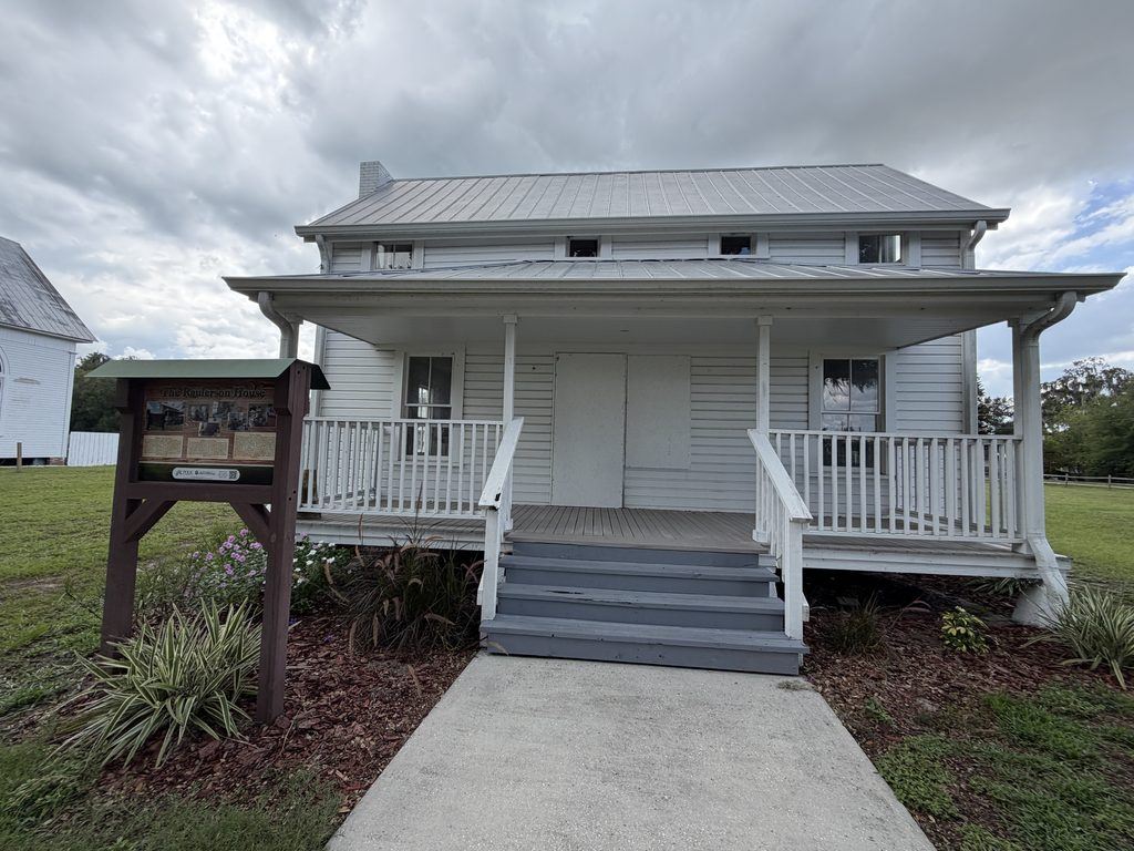 Front view of the historic white wooden Raulerson House at Homeland Heritage Park in Florida, featuring a broad covered porch, second-story windows, central staircase, and interpretive sign.