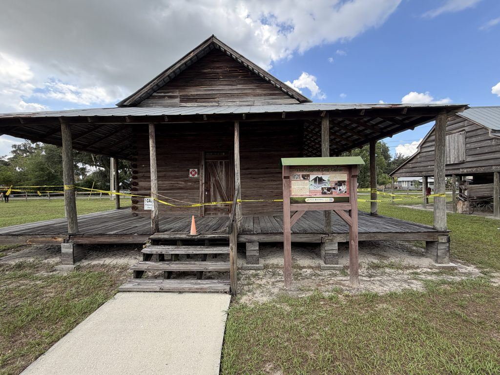Historic English family log cabin at Homeland Heritage Park in Polk County, Florida, featuring a rustic wooden exterior, wraparound porch, wooden steps, and interpretive sign.