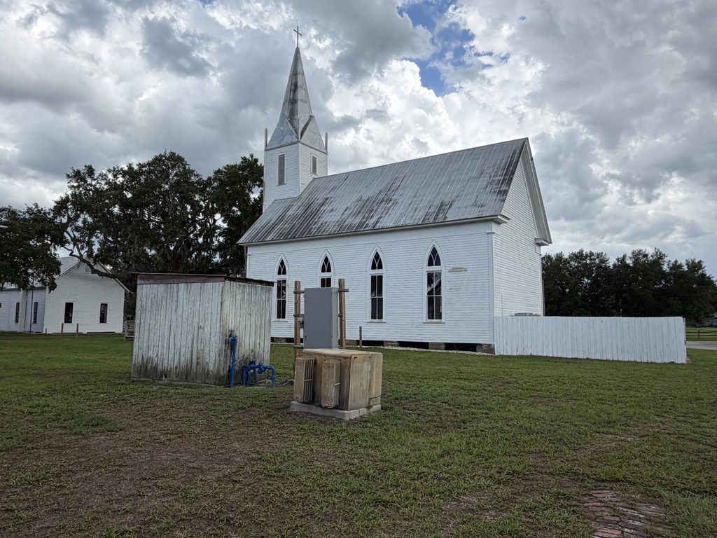 Rear and side view of the 1887 Old Homeland Methodist Church at Homeland Heritage Park in Polk County, Florida, showing its weathered metal roof, white wooden siding, pointed-arch windows, and tall steeple beneath dramatic clouds.