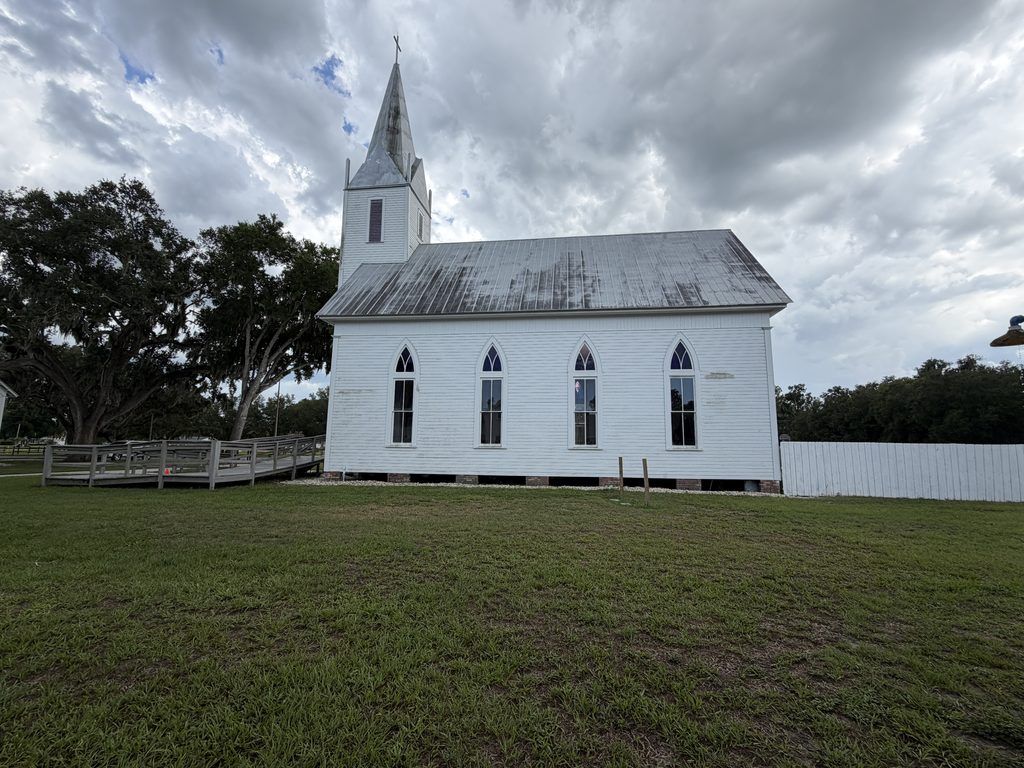 Side view of the 1887 Old Homeland Methodist Church at Homeland Heritage Park in Polk County, Florida, showing its white wooden siding, pointed-arch windows, metal roof, and tall steeple beneath cloudy skies.