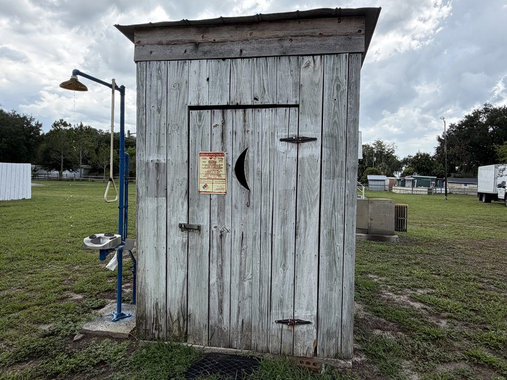 Weathered wooden outhouse with a crescent-moon cutout on its door, standing beside an outdoor emergency shower and eyewash station at Homeland Heritage Park in Polk County, Florida.