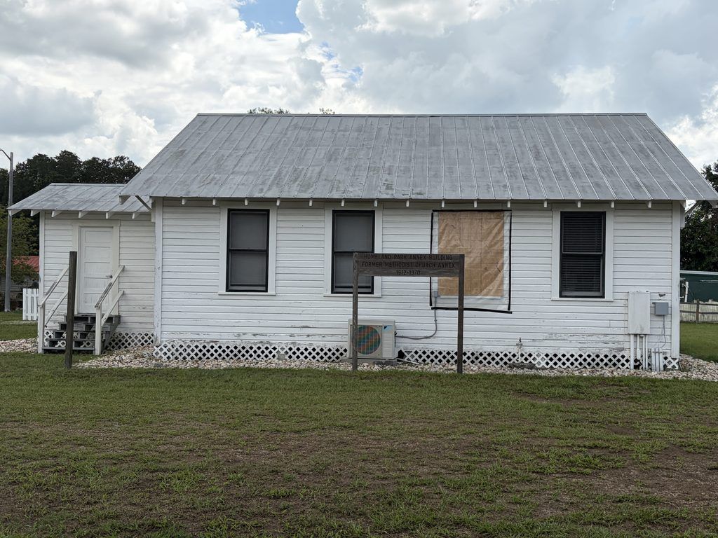 Historic white wooden Homeland Methodist Church parsonage at Homeland Heritage Park in Polk County, Florida, featuring a metal roof, covered porch, boarded windows, and a wooden identification sign beneath cloudy skies.
