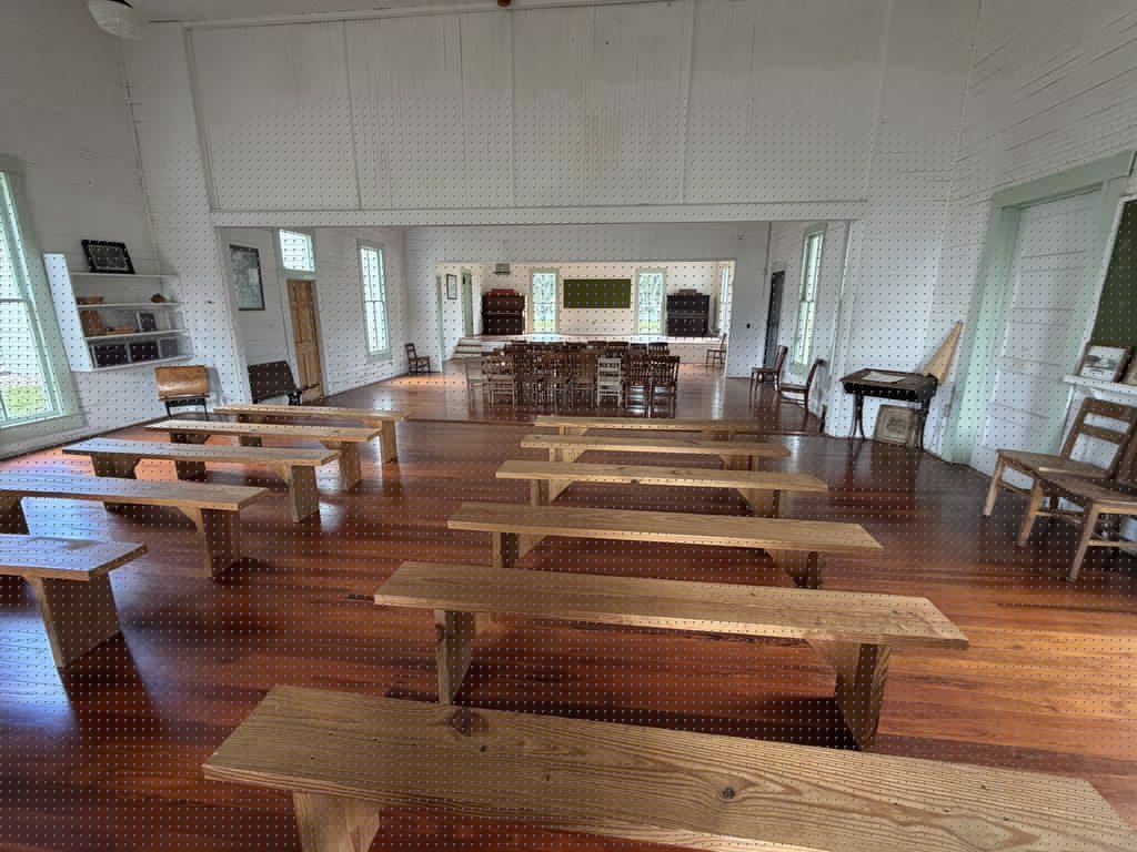 Interior of the historic Homeland School in Polk County, Florida, featuring wooden benches, polished floors, white plank walls, tall windows, and an adjoining classroom with antique desks and a chalkboard.